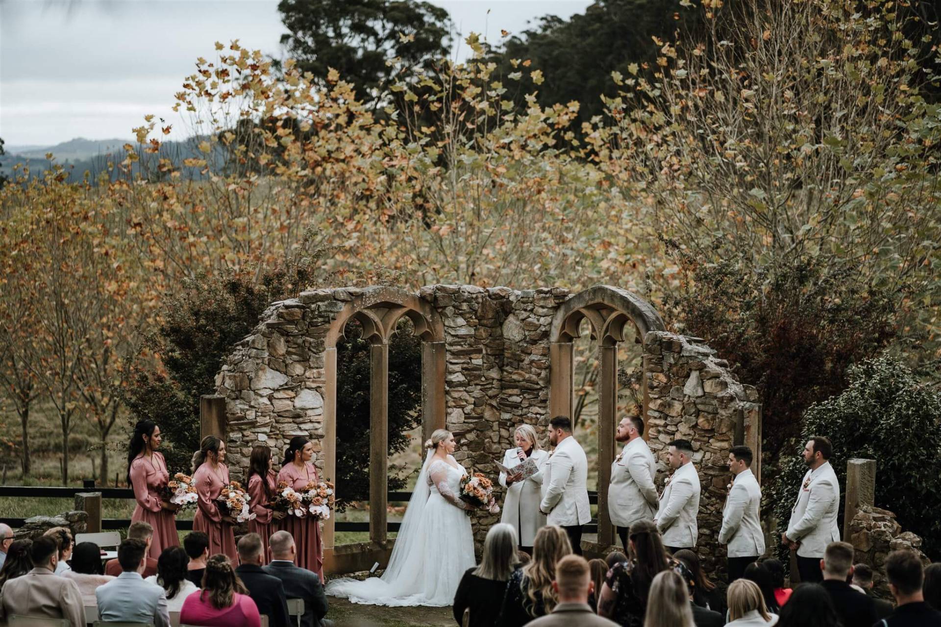 wedding ceremony vows at stone arch altar, surrounded by bridesmaids in rose dresses and groomsmen in white suits