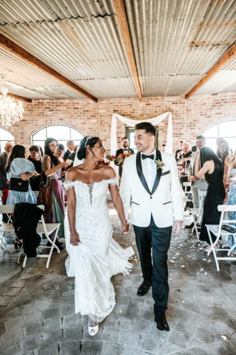 Newlyweds smiling as they walk through a rustic indoor venue with guests applauding