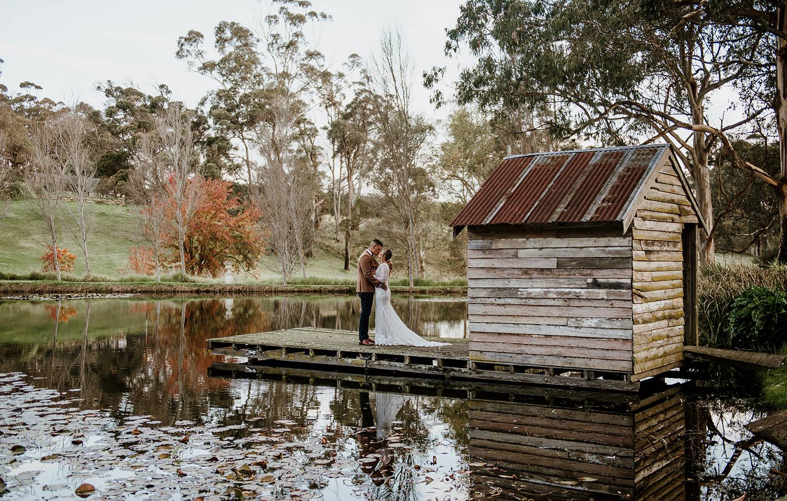 Couple sharing a tender moment on a rustic dock by a tranquil pond, with a quaint wooden hut and reflection in the water