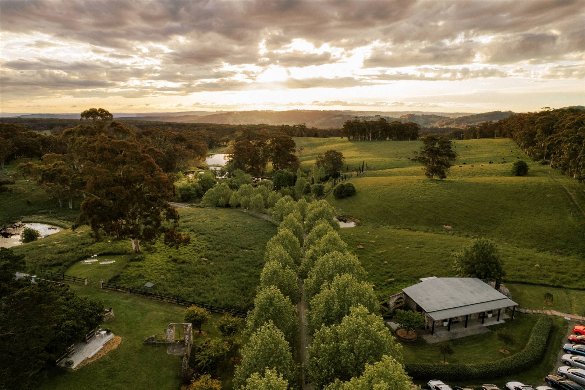 Aerial view of a lush wedding venue at sunset, with green fields, a pond, and a rustic building, under a dramatic sky