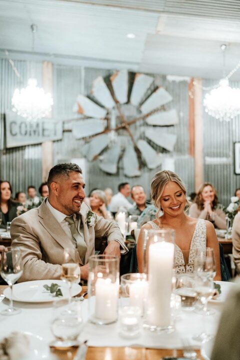 Newlywed couple seated at their wedding reception, smiling and enjoying the moment, with elegant candles and decor surrounding them