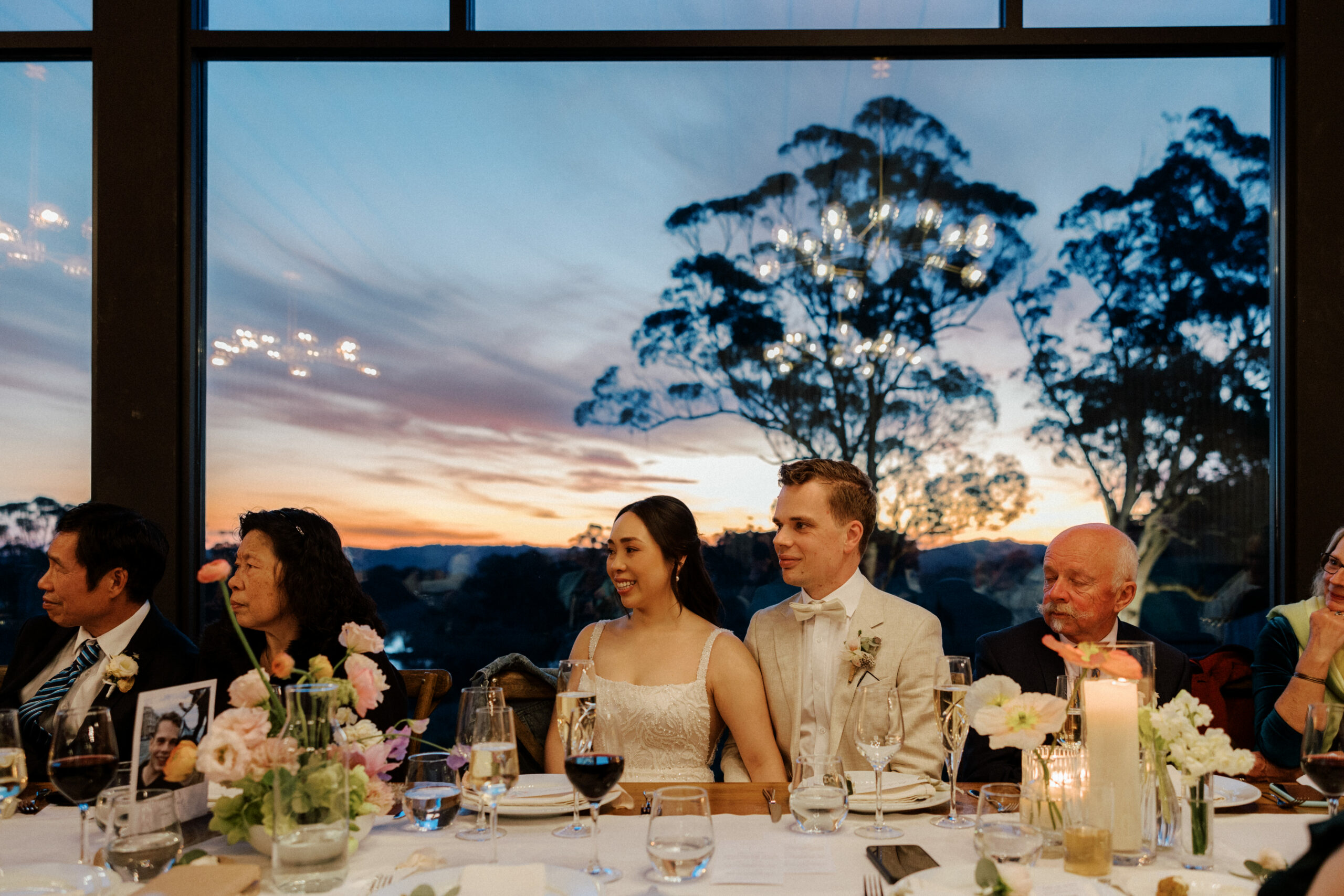 A bride and groom seated at a wedding reception table, looking out towards a large window that frames a stunning sunset, with guests and elegant table settings in the foreground