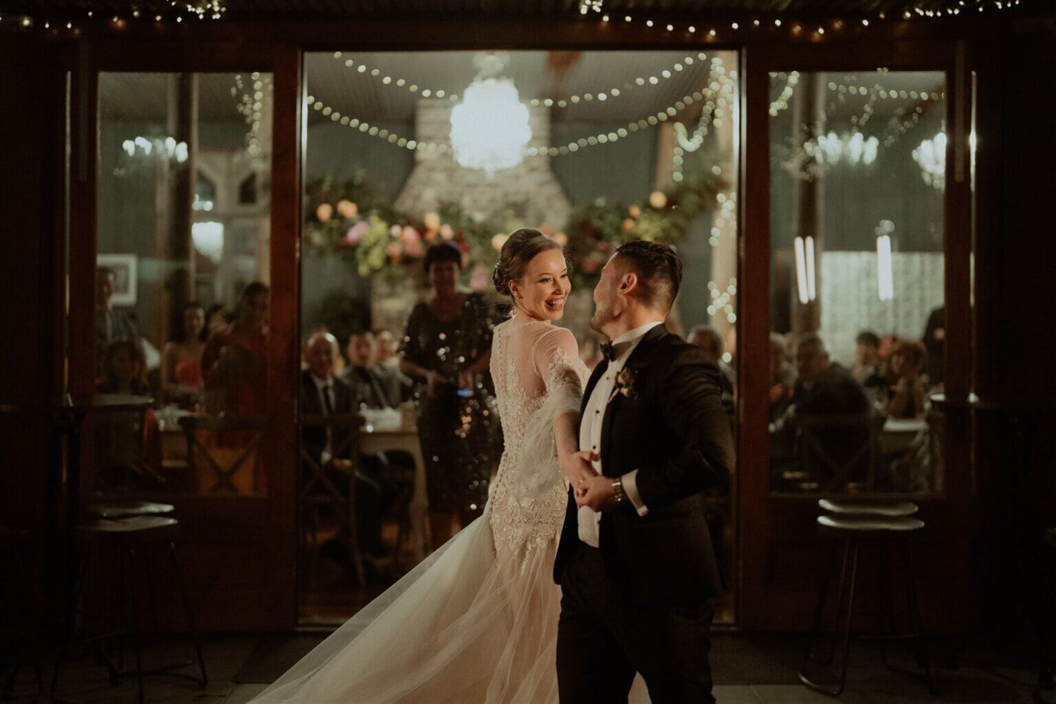 Newlyweds sharing their first dance in a rustic-chic venue with onlookers cheering, fairy lights twinkling in the background