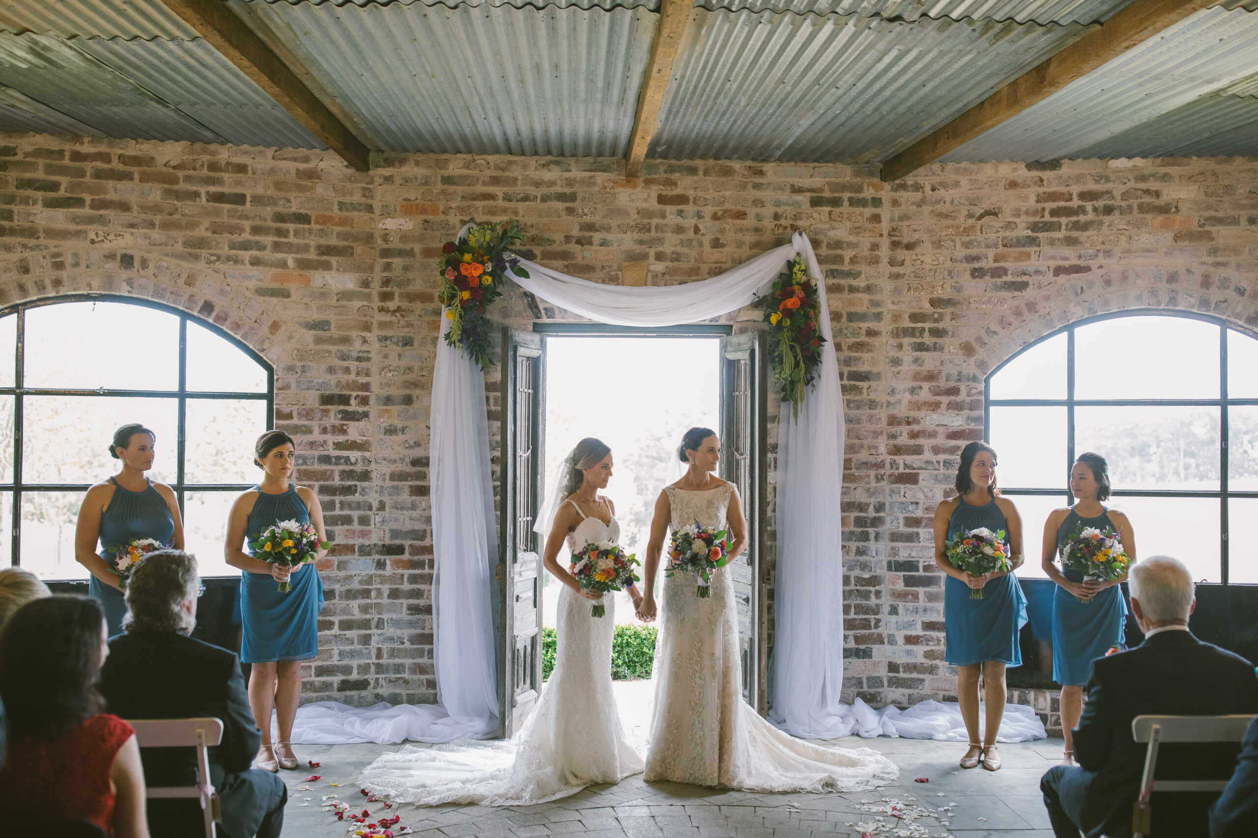 Brides and bridesmaids standing by the doorway in a rustic brick wedding venue, with bright floral arrangements