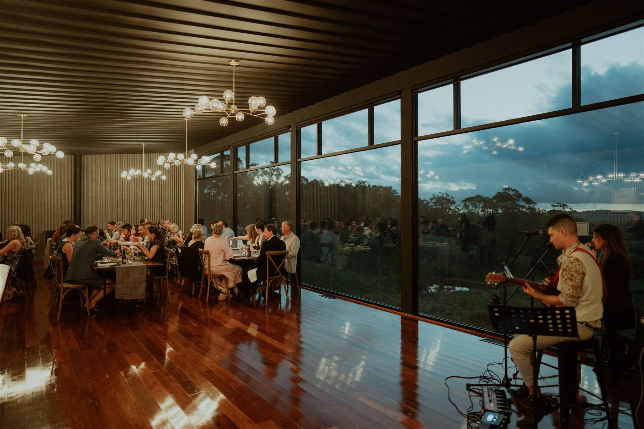An intimate indoor wedding venue with a rustic charm featuring guests dining under elegant globe chandeliers, as a musician plays guitar near large windows showcasing a dusky sky