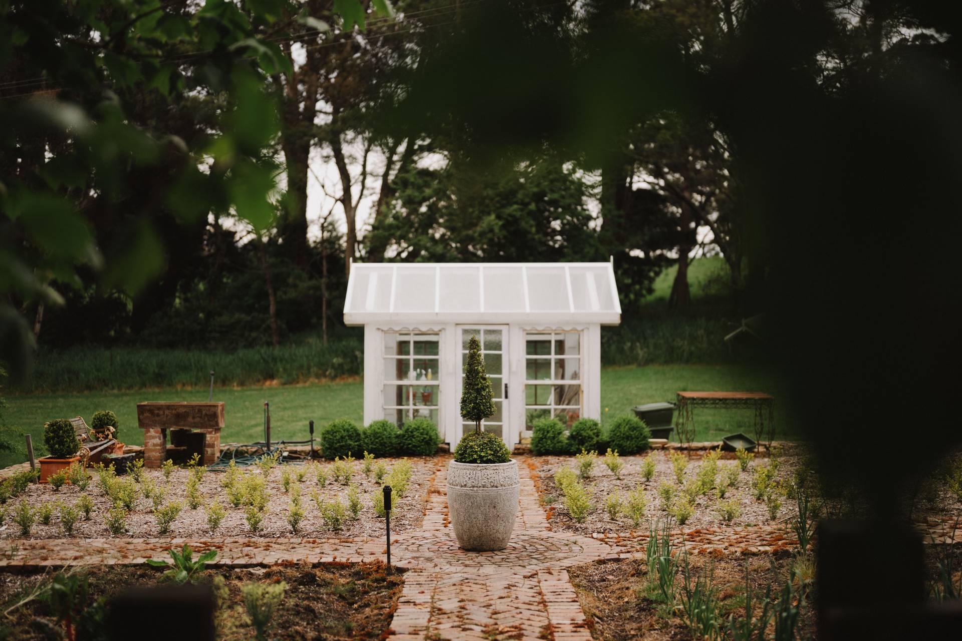 Quaint white greenhouse in a lush vegetable garden, with a brick path leading to it, in a serene rural setting