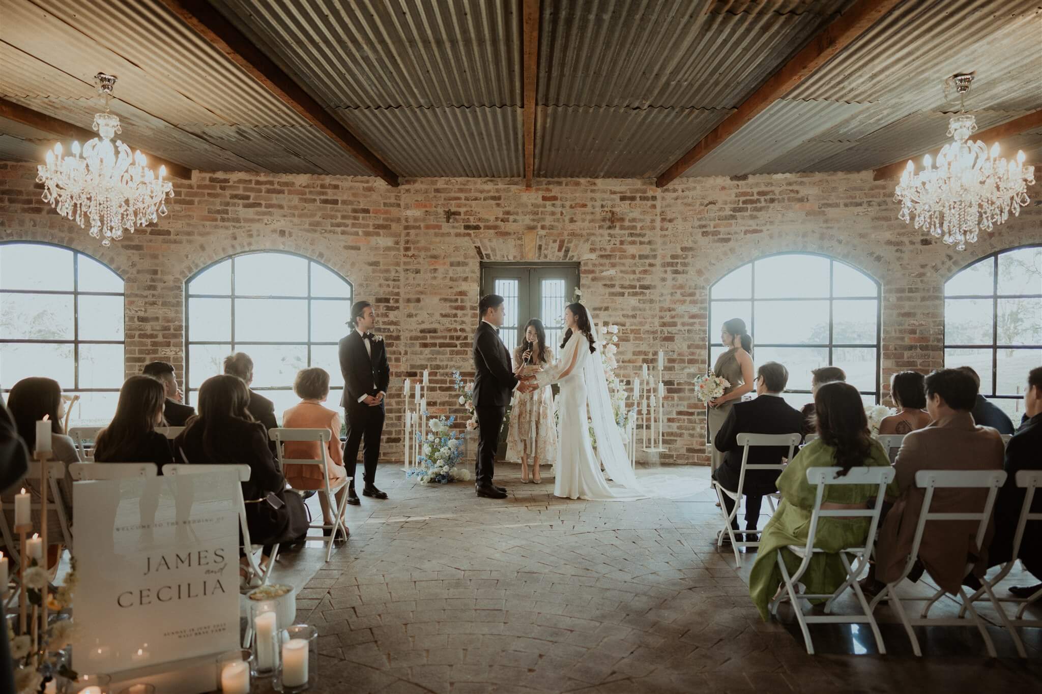A serene and elegant indoor wedding ceremony in a rustic venue with exposed brick walls and a high-beamed ceiling, as a couple stands at the altar with guests seated in rows, soft natural light streaming through arched windows