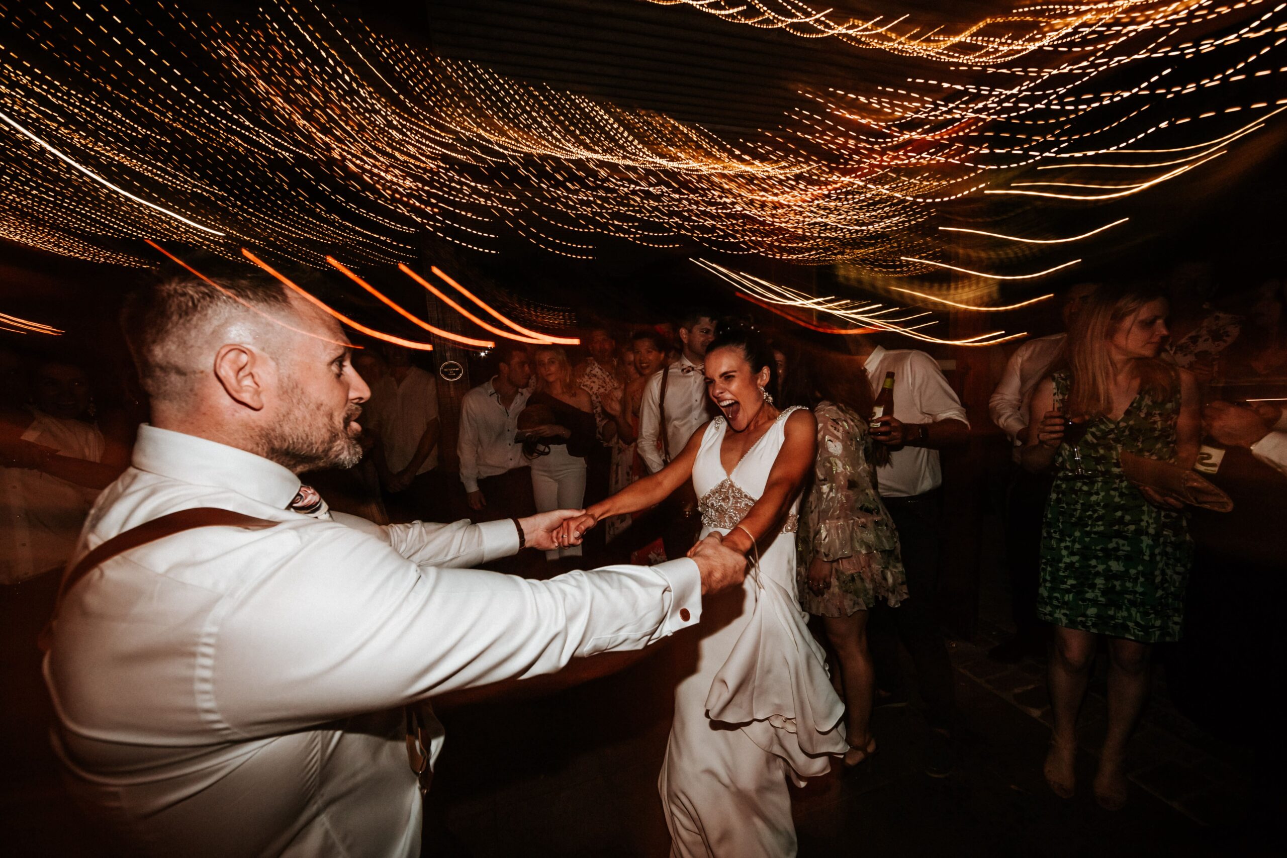 joyous dance floor moment at a wedding reception, with a bride in white spinning into the arms of a man in a suit, under a canopy of twinkling fairy lights creating a vibrant bokeh effect