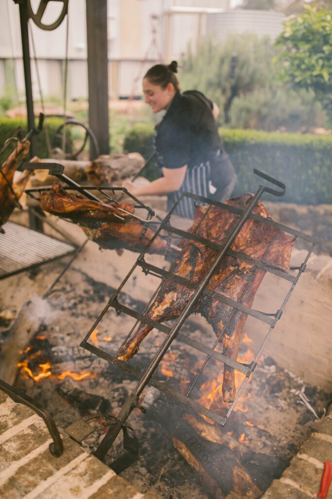 A chef attentively cooking large cuts of meat on a traditional open flame rotisserie, an authentic culinary experience at a wedding ceremony with smoke adding to the rustic atmosphere