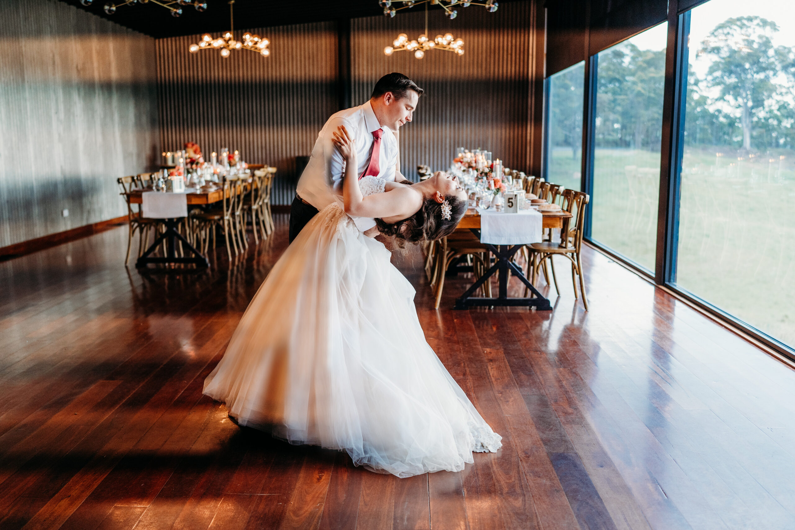 An intimate indoor wedding venue with a rustic charm featuring guests dining under elegant globe chandeliers, as a musician plays guitar near large windows showcasing a dusky sky